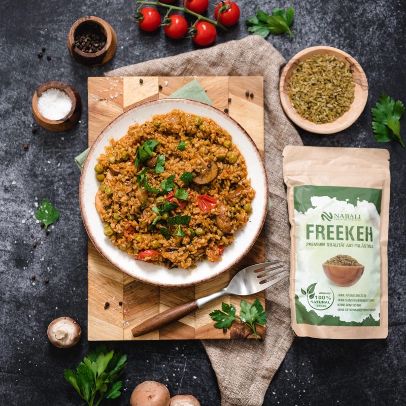 Ein Teller Freekeh mit Gemüse und Kräutern, umgeben von frischen Zutaten und einer Packung Freekeh.