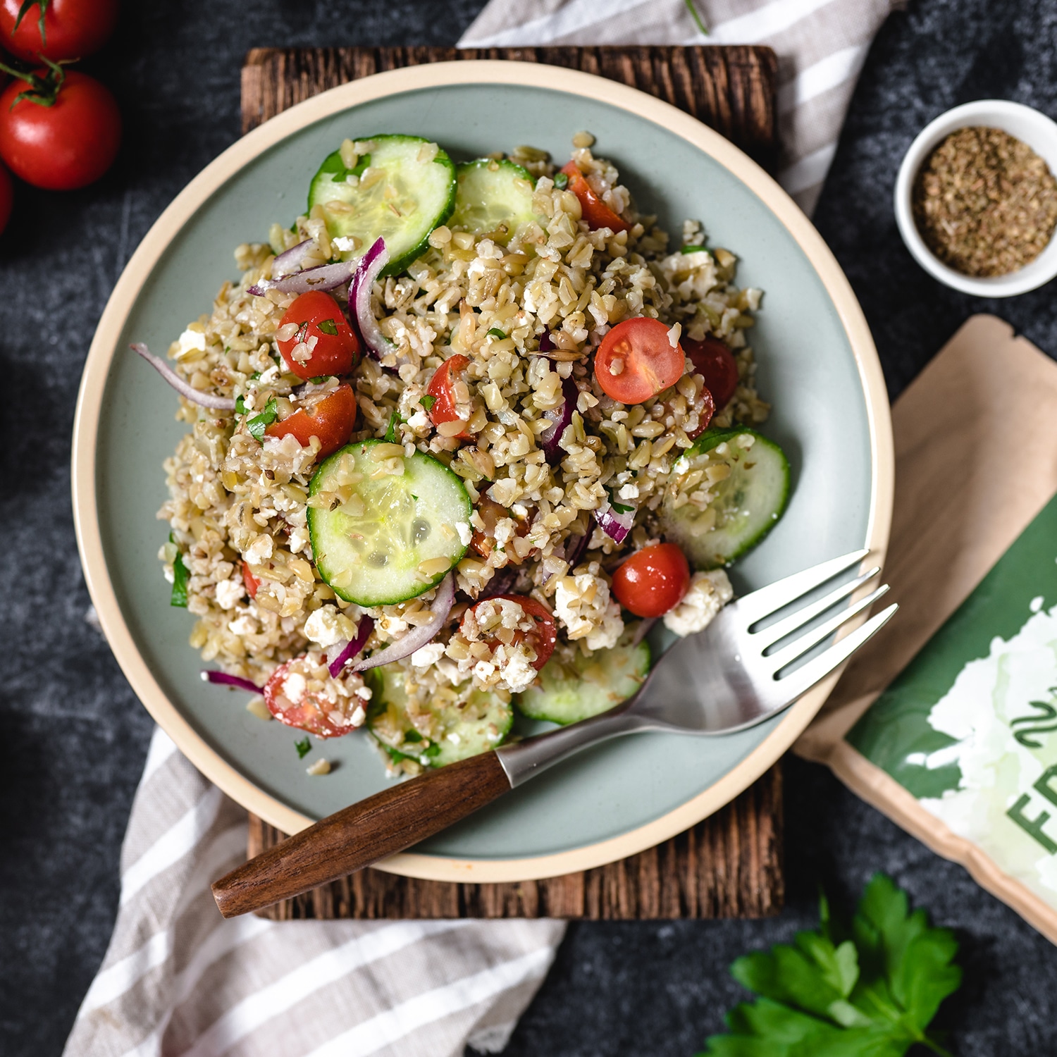 Ein frischer Freekeh-Salat mit Gurkenscheiben, Kirschtomaten, roten Zwiebeln und Feta-Käse, auf einem rustikalen Holzbrett serviert.