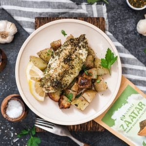 Ein Teller mit Lachs, der mit Zaatar gewürzt ist, serviert auf einem Bett aus goldbraun gebratenen Ofenkartoffeln. Im Hintergrund sind Knoblauch, Salz und eine Packung Zaatar-Gewürz zu sehen.